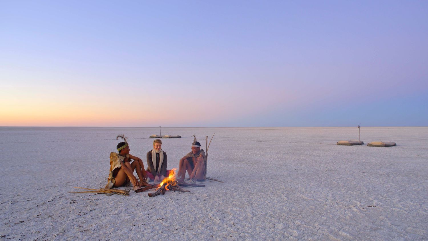 Three individuals sitting by a campfire on a vast salt flat at sunset, with a soft gradient of purple and orange in the sky. The landscape is flat and barren, with a few stones visible in the background.