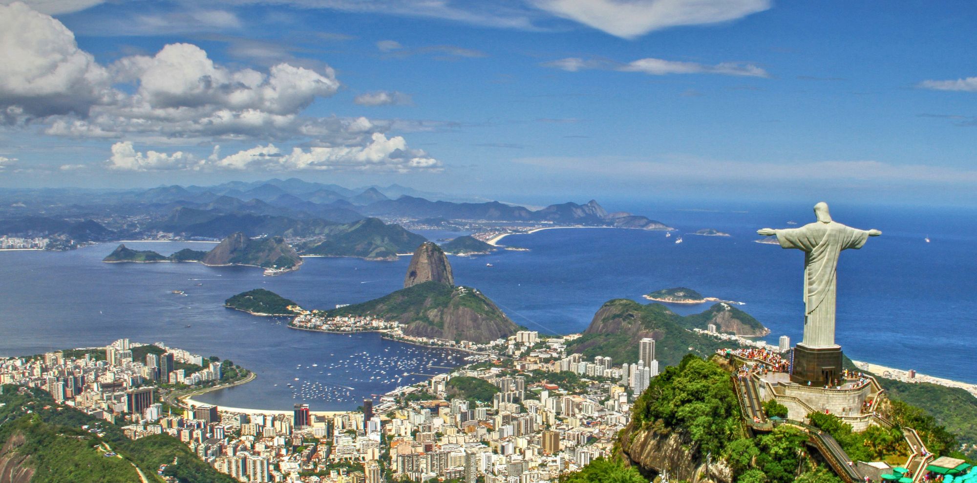 A panoramic view of Rio de Janeiro featuring the iconic Christ the Redeemer statue on Corcovado Mountain. The cityscape includes lush mountains, coastal beaches, and the bay with boats, under a blue sky with scattered clouds.