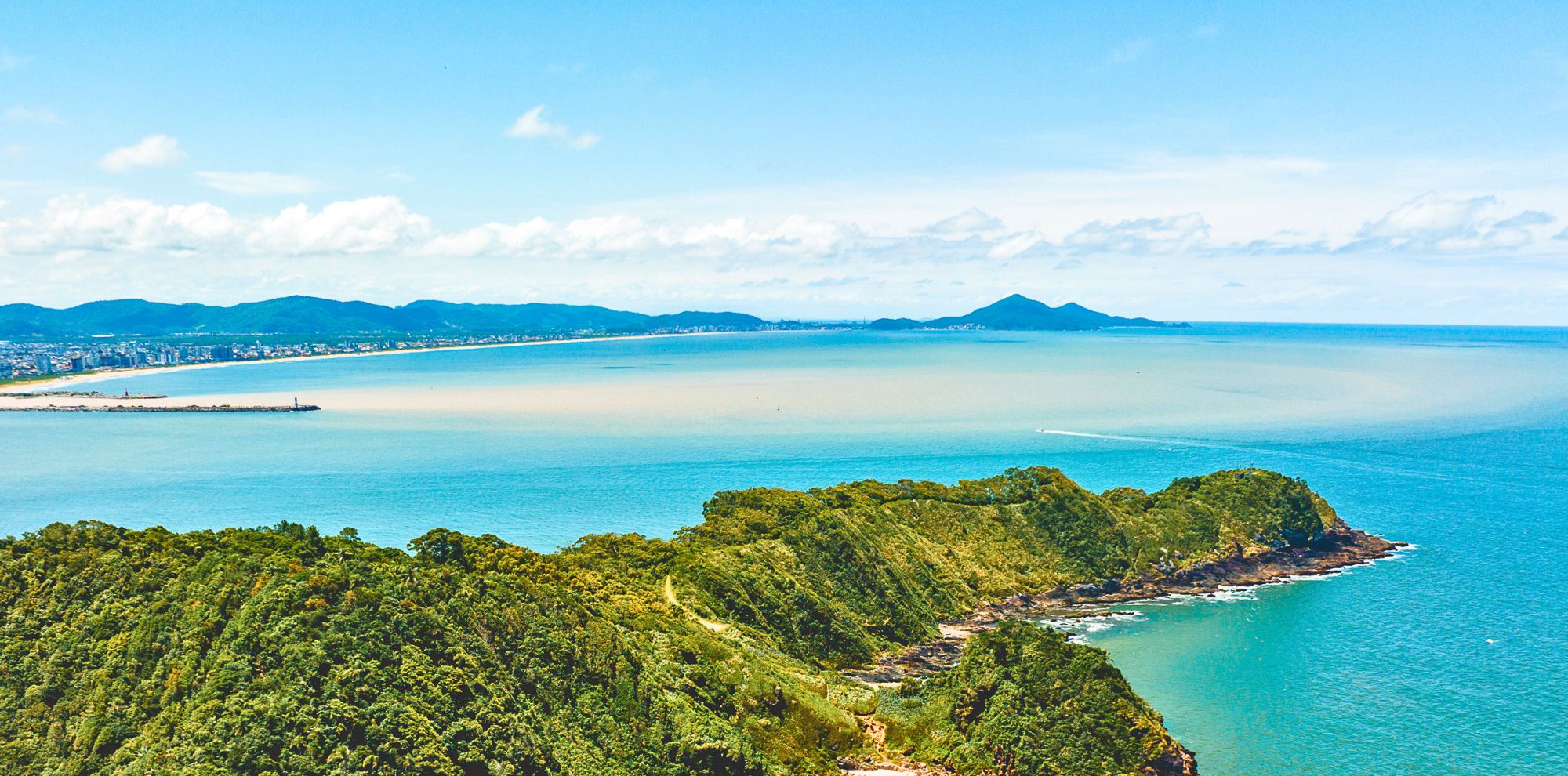 A panoramic view of a coastline featuring lush green hills, sandy beaches, and a serene blue sea under a bright sky with scattered clouds. In the distance, a mountain range is visible across the water.