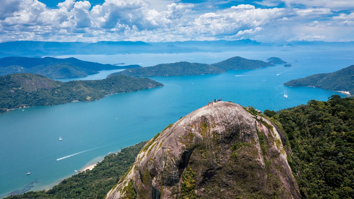 Aerial view of a rocky summit with a group of hikers, surrounded by lush greenery and a breathtaking seascape featuring numerous islands and sailboats in the sparkling blue water under a partly cloudy sky.