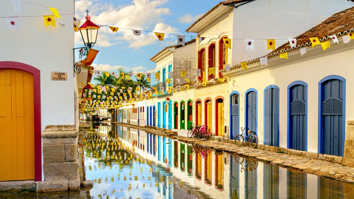 A picturesque street in a vibrant town, featuring colorful buildings with blue, yellow, and white facades. The scene is reflected in a calm canal, adorned with festive bunting and lanterns, and bicycles parked along the walkway.