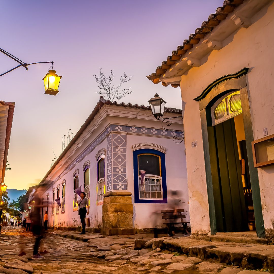 Historic cobblestone street lined with charming colonial buildings, featuring a warm sunset glow in the background. Lanterns illuminate the scene, while a person stands by a doorway, adding to the quaint atmosphere.
