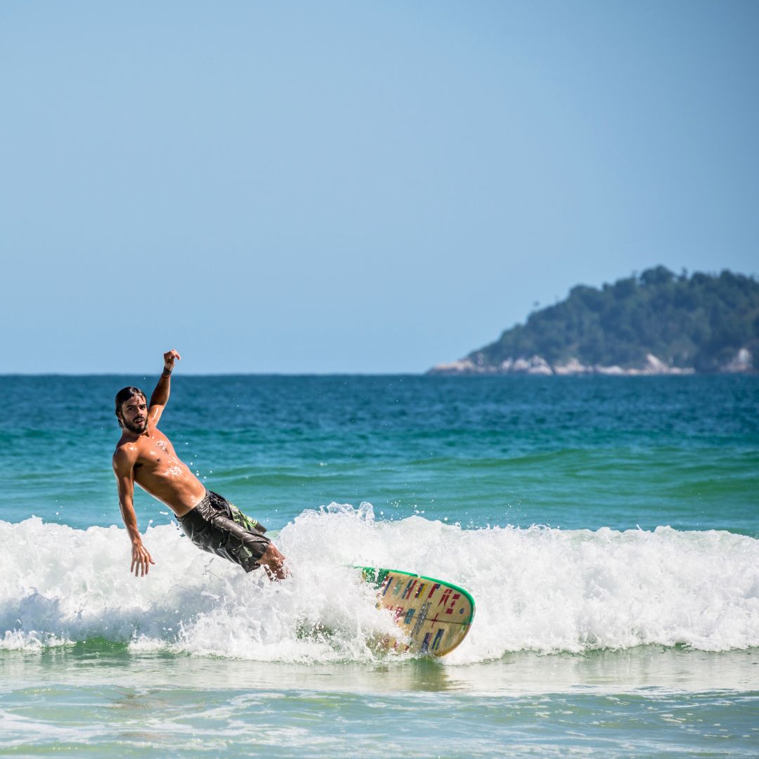 A man is surfing on a wave at the beach, balancing on his surfboard with one arm raised. The ocean is blue, and a green island is visible in the background.