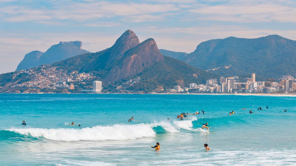 A vibrant beach scene with surfers riding the waves in the foreground. Gorgeous mountains rise behind a coastal city, with sunlit buildings and lush greenery. The sky is partly cloudy, adding a tranquil atmosphere to the lively beach activity.