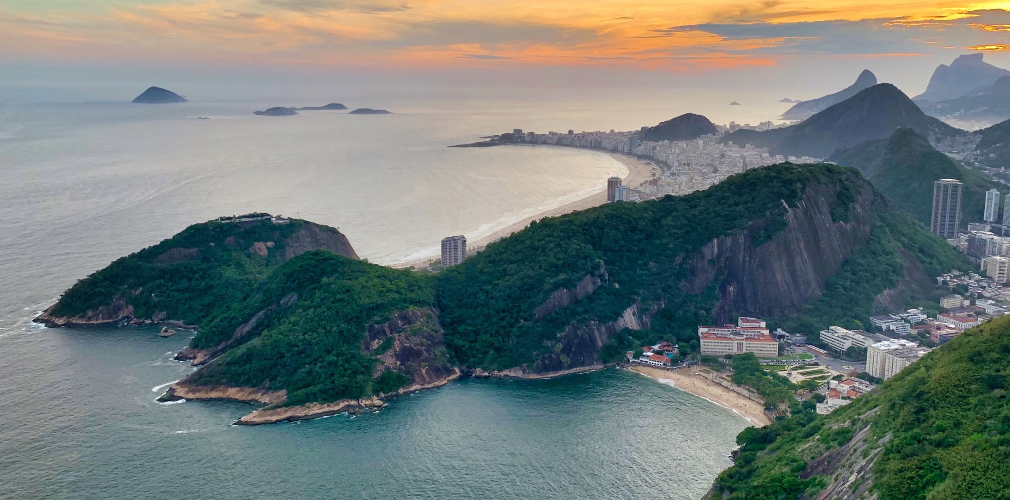 A stunning coastal view of a green mountainous landscape meeting the ocean at sunset, with a city skyline in the distance and islands dotting the horizon.