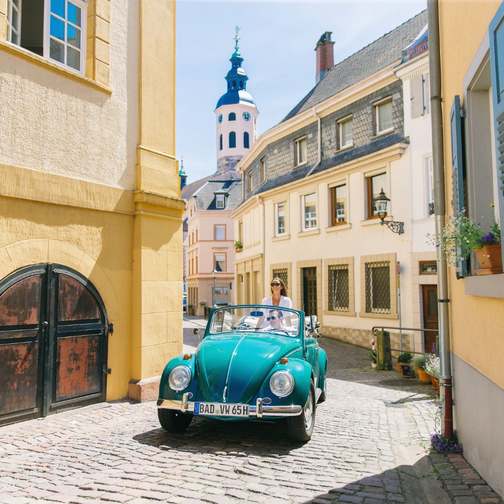 A blue vintage convertible car parked on a cobblestone street with a historic tower in the background. A person sits in the driver's seat, surrounded by charming buildings and warm sunlight.