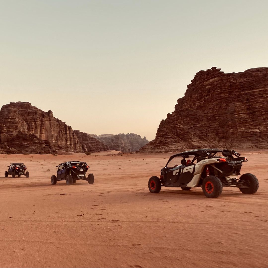Three all-terrain vehicles racing across a sandy desert landscape, with towering rock formations in the background. The sun is setting, casting a warm glow over the scene.