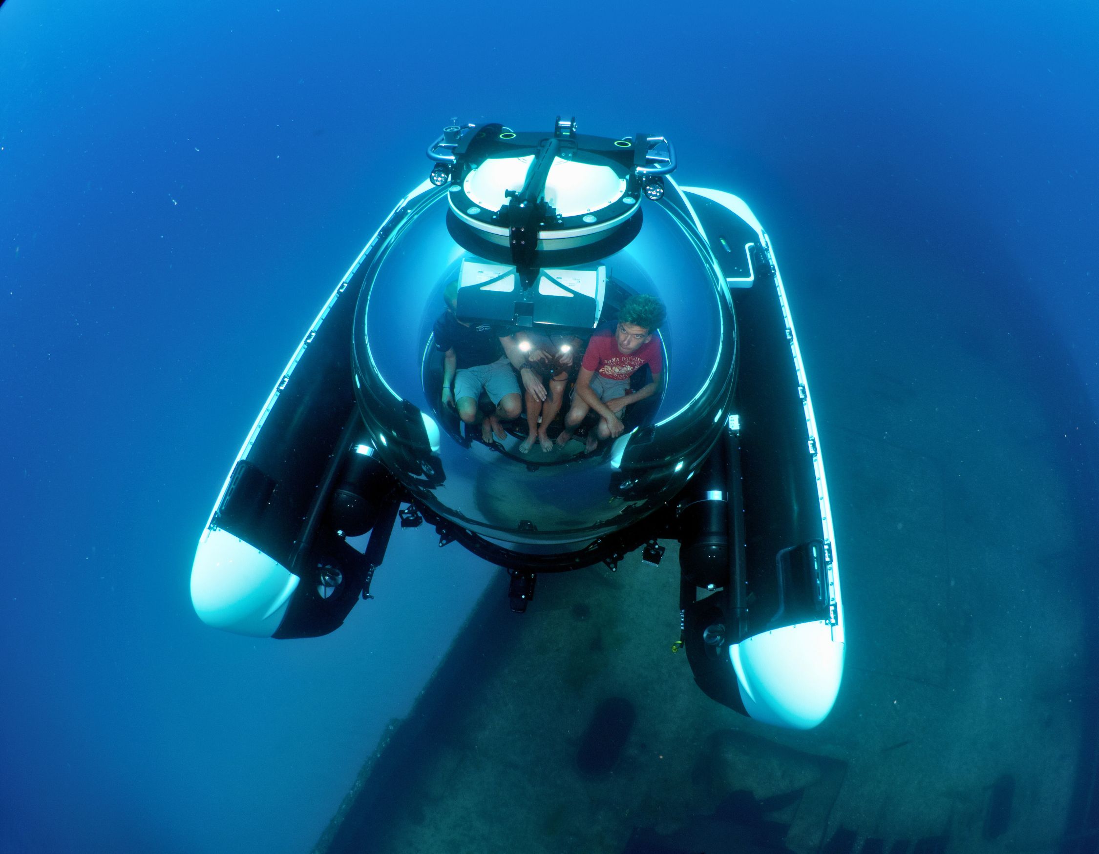 A submersible with three passengers is underwater, exploring a sunken ship. The clear dome allows visibility of the occupants and the surrounding blue ocean.