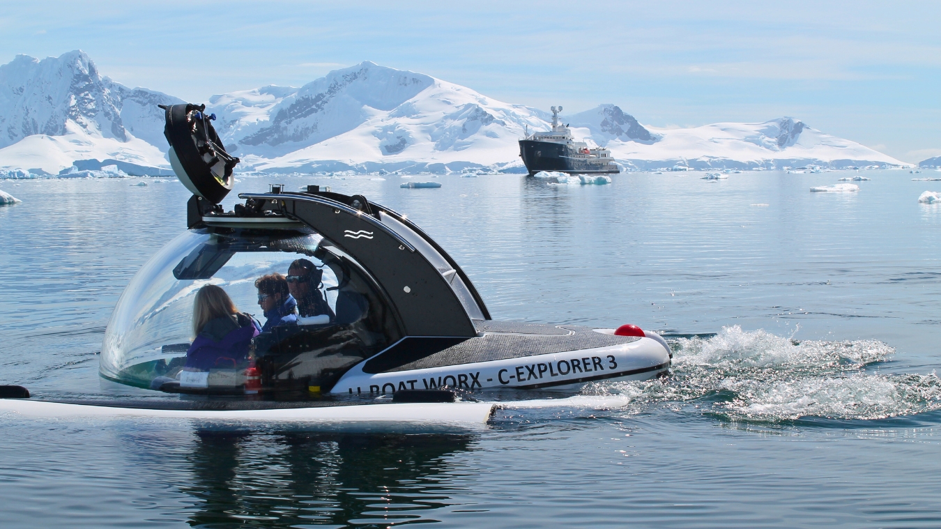 A transparent submersible, C-Explorer 3, glides through calm waters surrounded by icebergs, with snowy mountains in the background. Two passengers are seated in the submersible, exploring the Antarctic landscape while a ship is visible in the distance.