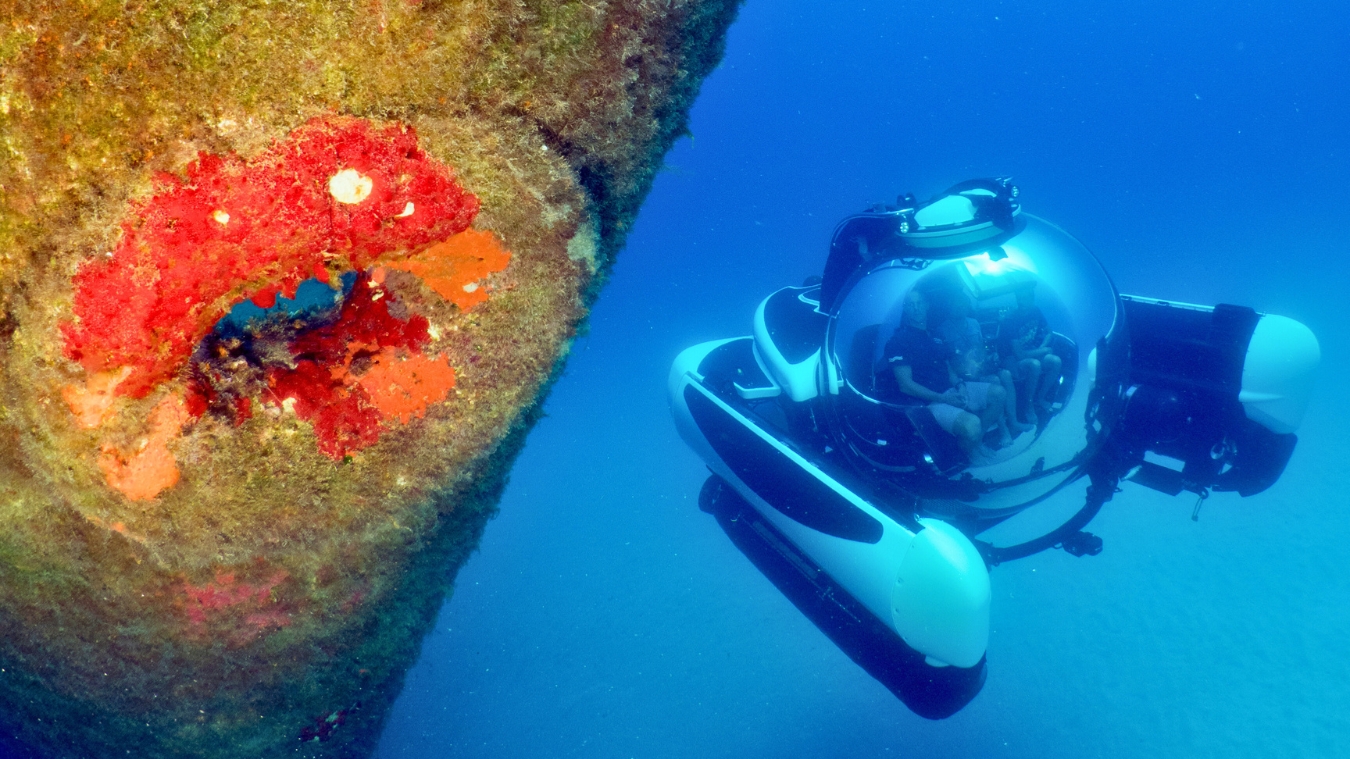 A submersible explores a submerged structure covered in vibrant red coral, with the ocean's blue depths in the background.