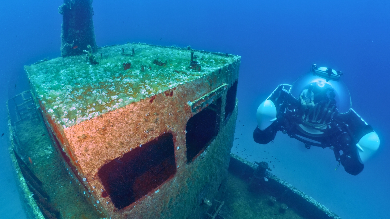 A submersible exploring a sunken shipwreck, with coral and marine life covering the vessel's structure in clear blue water.