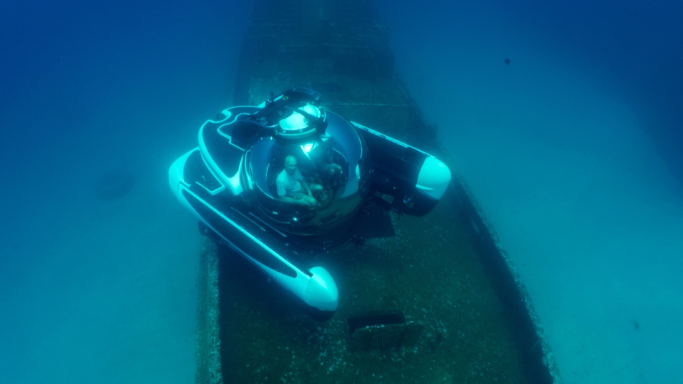 A submariner operating a deep-sea submersible above the remains of a submerged shipwreck, surrounded by clear blue water. The submersible features a round cockpit and sleek design, highlighting its exploration capabilities.