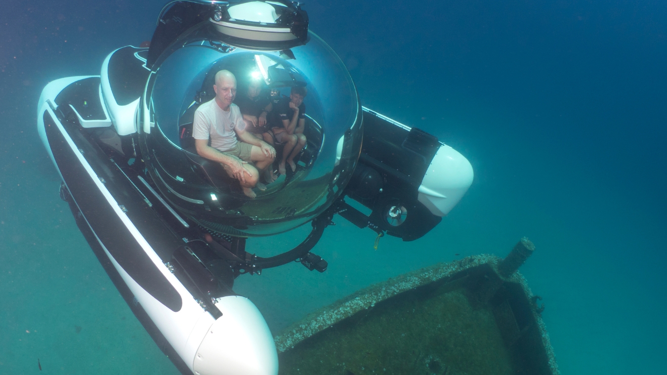 A group of three people seated inside a submersible, with a large, rounded glass canopy, exploring a sunken ship underwater. The clear blue water surrounds them, and the silhouette of the shipwreck is visible below.