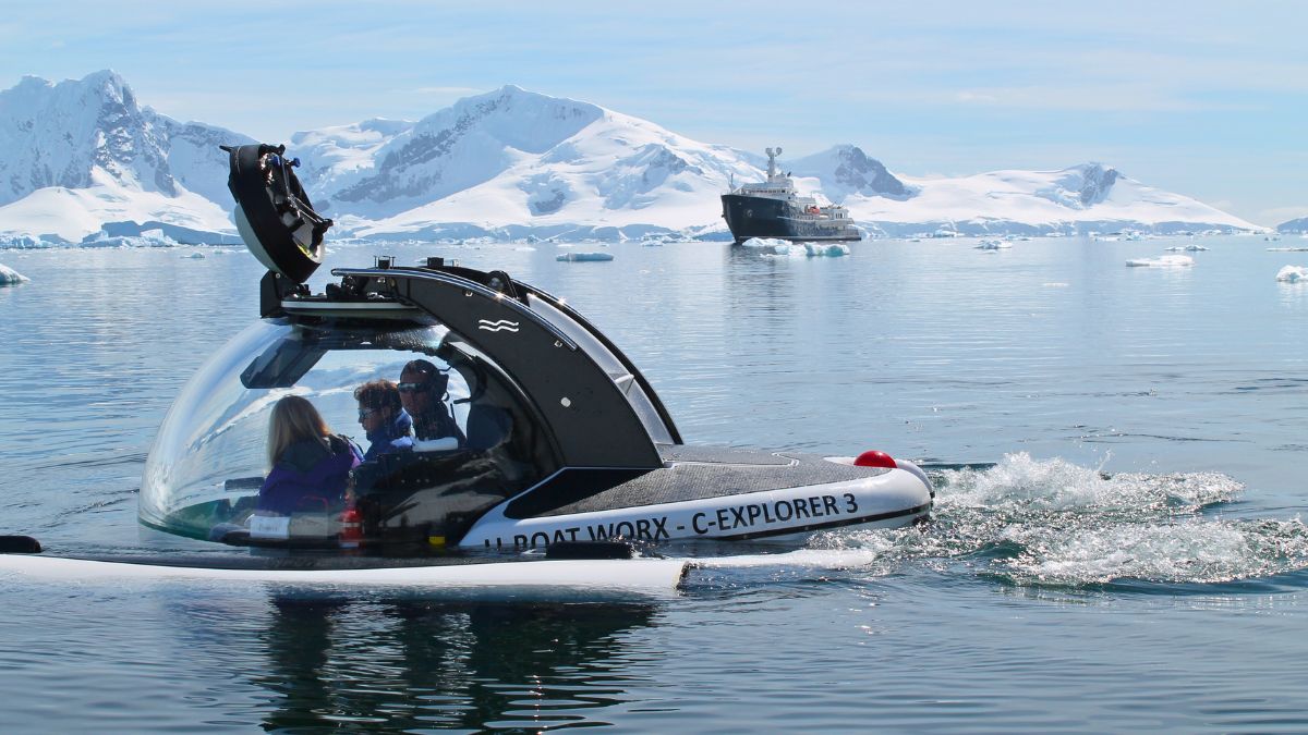 A transparent submersible with two people inside glides through calm icy waters, surrounded by snow-capped mountains and icebergs. A research vessel is visible in the distance.