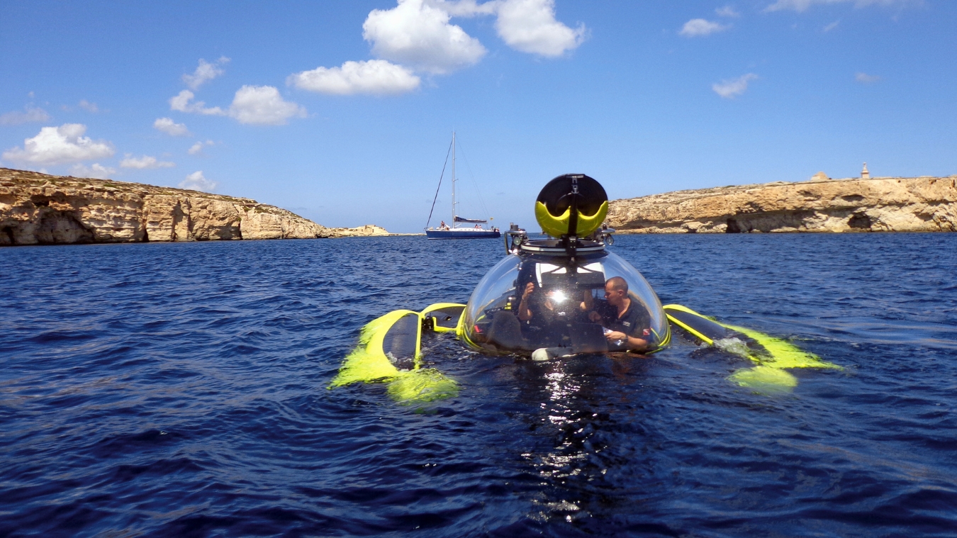 A bright yellow submarine is floating in clear blue water near rocky cliffs. Two people can be seen inside, and a sailboat is moored in the background under a partly cloudy sky.