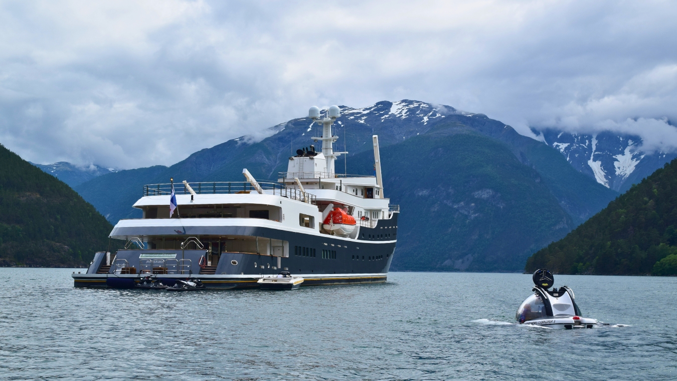A large luxury yacht anchored in a serene fjord, with mountains in the background and a small submarine nearby. The sky is overcast, enhancing the tranquil atmosphere.