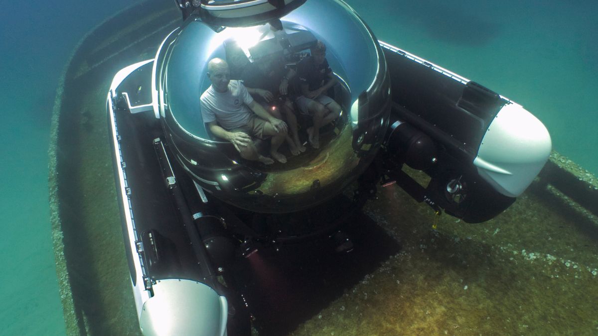 A submersible exploring a sunken shipwreck underwater, with two people inside visible through a large, round viewing dome. The ocean floor is visible, along with marine life and the structure of the ship.
