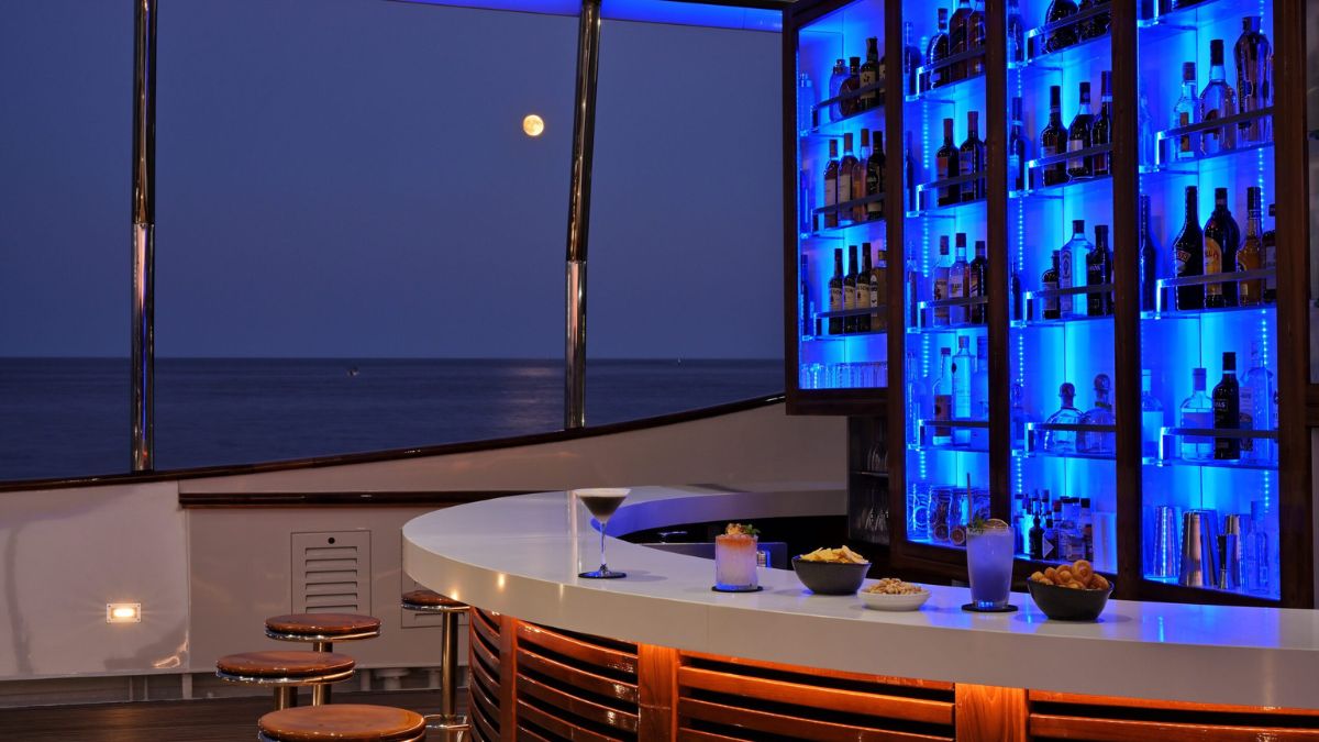 A stylish bar area with a sleek white counter, featuring a variety of illuminated bottles on shelves. The scene is set against a twilight ocean view with a full moon visible in the sky. Snacks are placed on the counter, alongside cocktails.