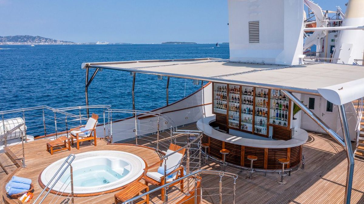 A luxurious yacht deck featuring a hot tub and a circular bar stocked with various bottles, surrounded by wooden lounge chairs. Soft blue ocean waters stretch out in the background under a clear sky.