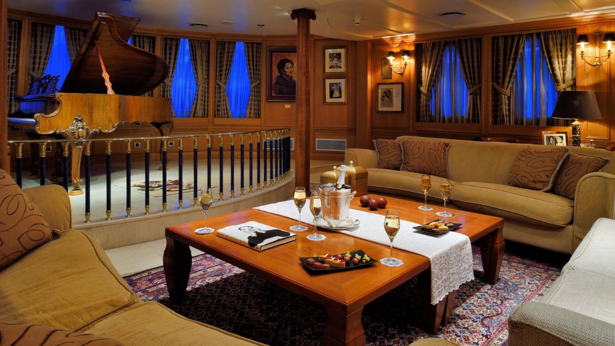 Elegant lounge area featuring a grand piano, plush seating, a coffee table with a selection of snacks and drinks, and warm wooden accents. Soft lighting highlights the rich decor and artwork on the walls.