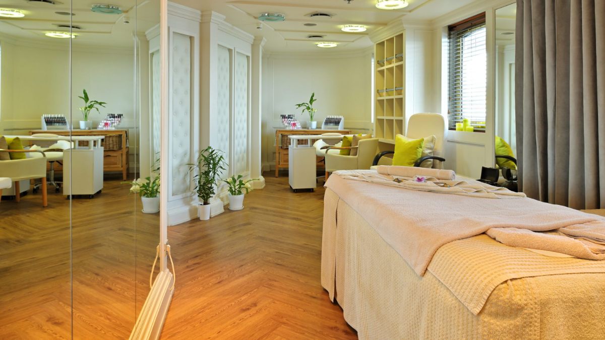 A serene spa interior featuring a treatment table with a light beige cover, surrounded by plants and natural light. Soft seating area with green cushions and a mirrored wall reflecting the calm atmosphere.
