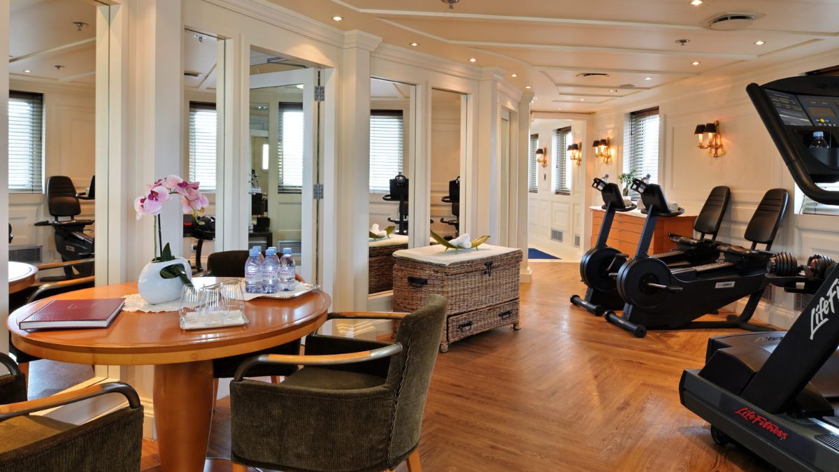 A well-lit exercise room featuring gym equipment like stationary bicycles and a treadmill, with a cozy seating area including a round wooden table, chairs, and a decorative plant. Bottled water and a menu are on the table.