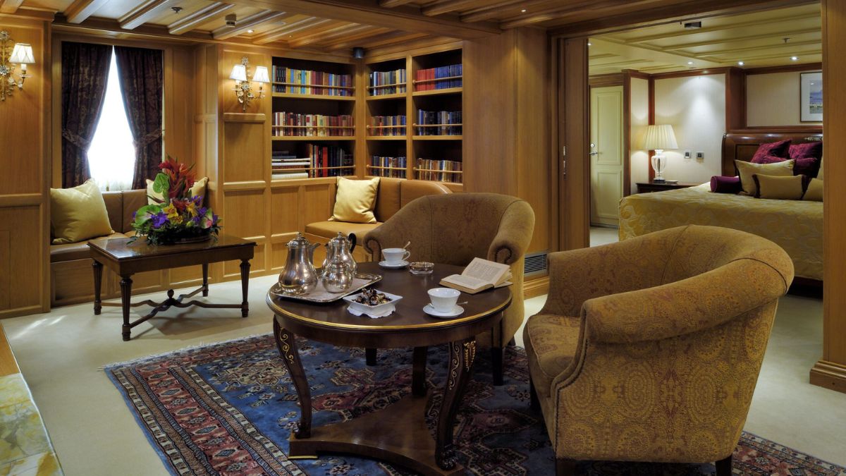 A cozy, well-appointed room features plush armchairs, a coffee table set with tea and pastries, a bookshelf filled with books, and elegant decor. Soft lighting enhances the warm, inviting atmosphere.