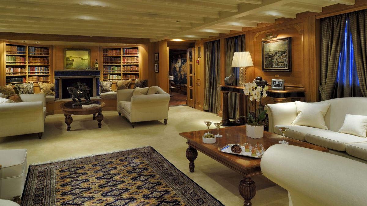 A cozy, elegantly furnished living room featuring two cream-colored sofas, a wooden coffee table with snacks, and a large bookcase filled with books. Soft lighting from a lamp illuminates the space, which is decorated with artwork and a large area rug. Dark curtains frame the windows, providing a warm atmosphere.