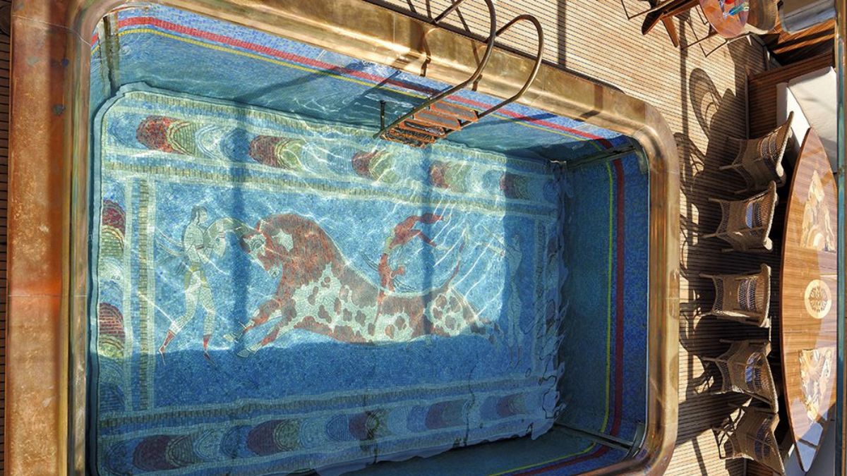 Aerial view of a decorative swimming pool featuring a mosaic design of a bull on the bottom, surrounded by blue water, with a ladder on one side and seating area nearby.
