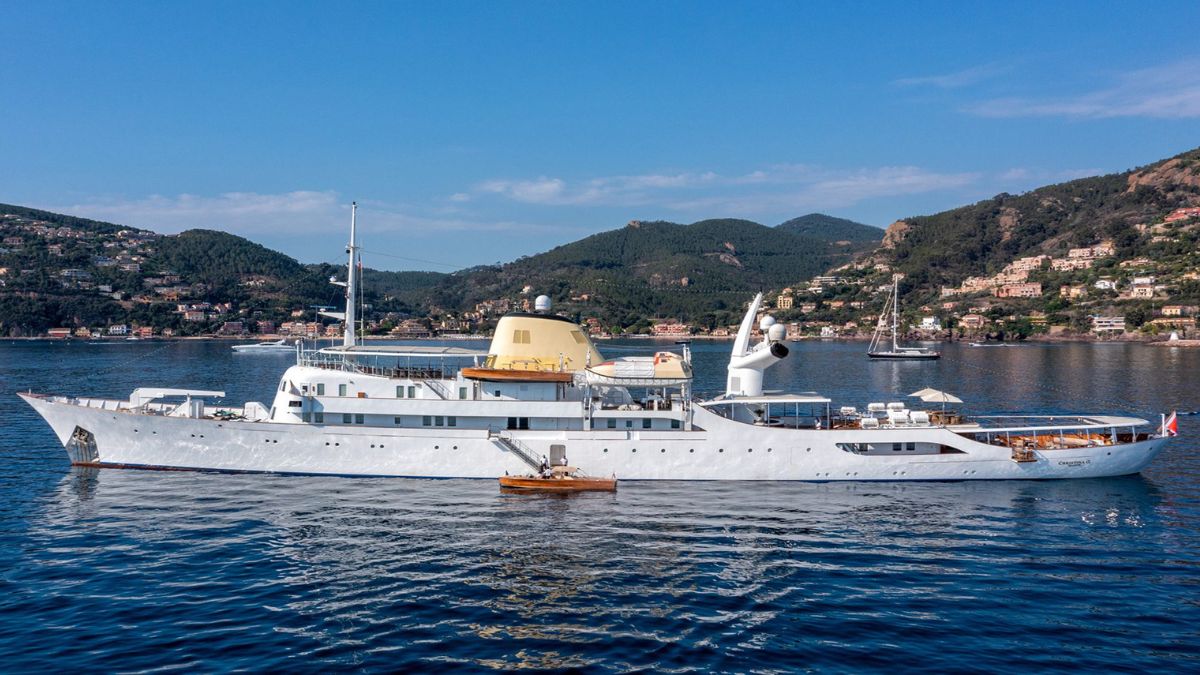 A luxurious yacht anchored in calm waters, surrounded by lush green hills and a clear blue sky. The yacht features a sleek design with a prominent yellow dome and multiple decks. Smaller boats are visible nearby.