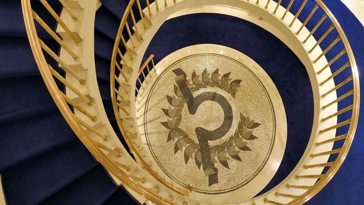 A spiral staircase with a golden railing. The floor features a decorative mosaic design surrounded by a blue carpet.