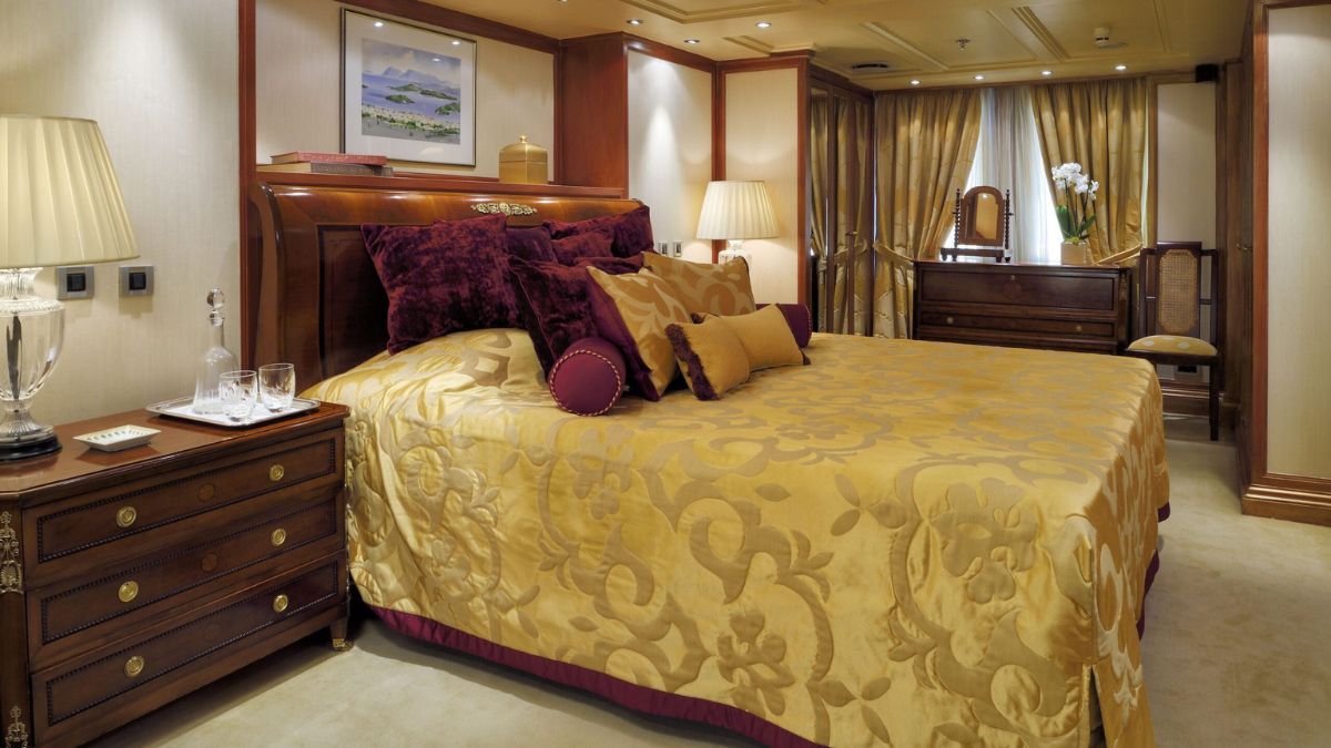 Luxurious bedroom with a king-size bed adorned with golden bedding and decorative pillows. Elegant wooden furniture, including a bedside table and dresser, complements the warm, rich tones of the decor. Soft lighting from lamps adds to the cozy ambiance. A piece of art hangs above the bed, and curtains frame the window, enhancing the opulent atmosphere.