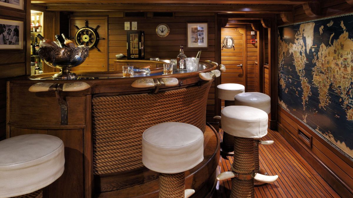 A wooden bar area featuring a rounded counter with a nautical theme, several round stools with white cushions, and a vintage world map on the wall. Nautical decor elements, including a ship's wheel and various photographs, are present. The warm wooden tones create a cozy atmosphere.