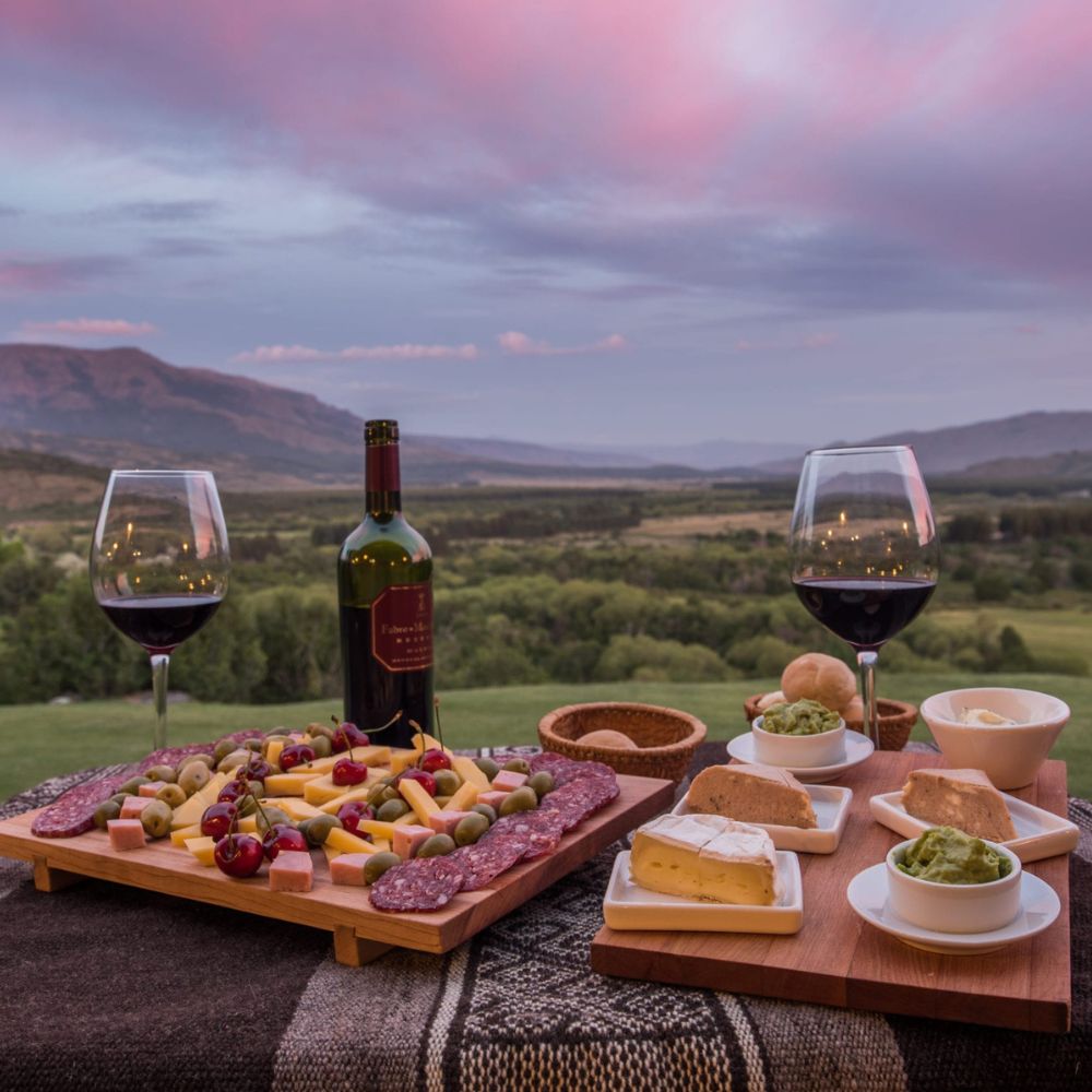 A beautifully arranged platter with a variety of cheeses, cured meats, olives, and grapes, accompanied by two glasses of red wine and a bottle. The scene is set against a picturesque landscape with rolling hills and a colorful sunset.