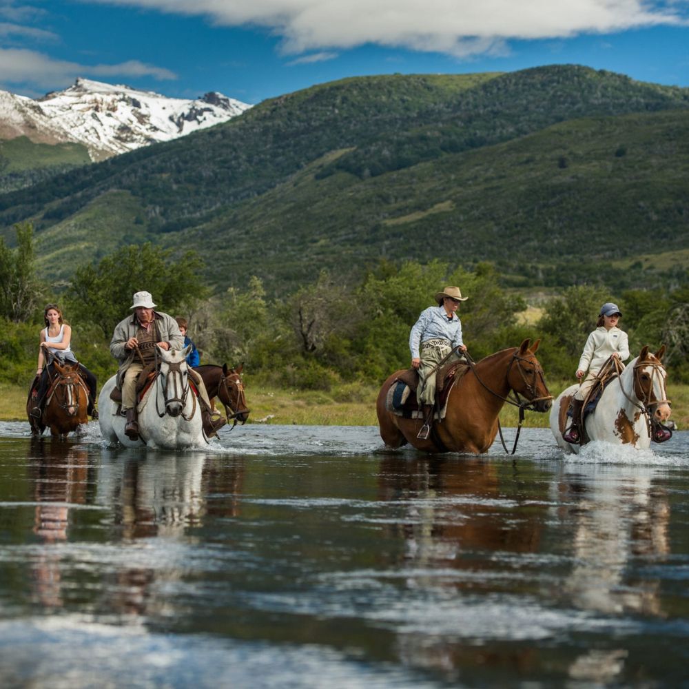 Four horseback riders cross a shallow river, surrounded by lush greenery and mountainous terrain in the background. Snow-capped peaks are visible under a blue sky with scattered clouds.