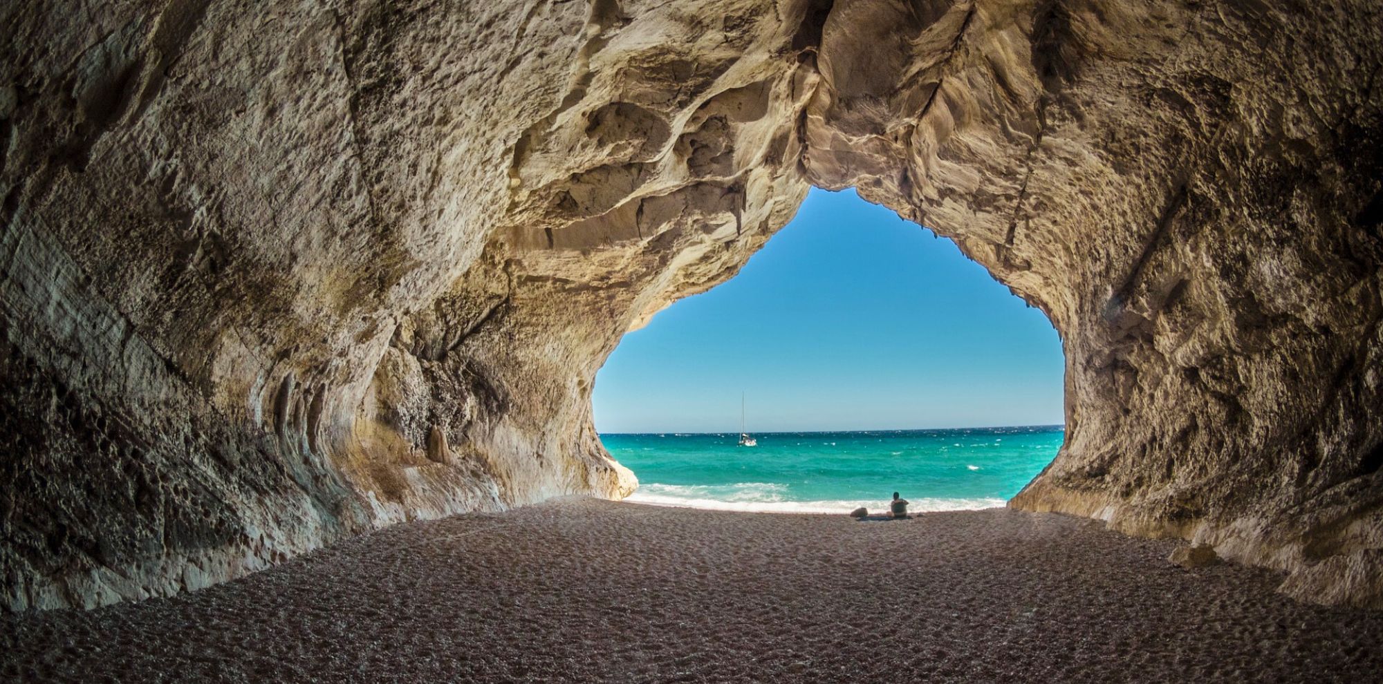 A stunning coastal cave framing a picturesque view of the vibrant blue ocean and a sailboat on the horizon, with a sandy beach in the foreground.