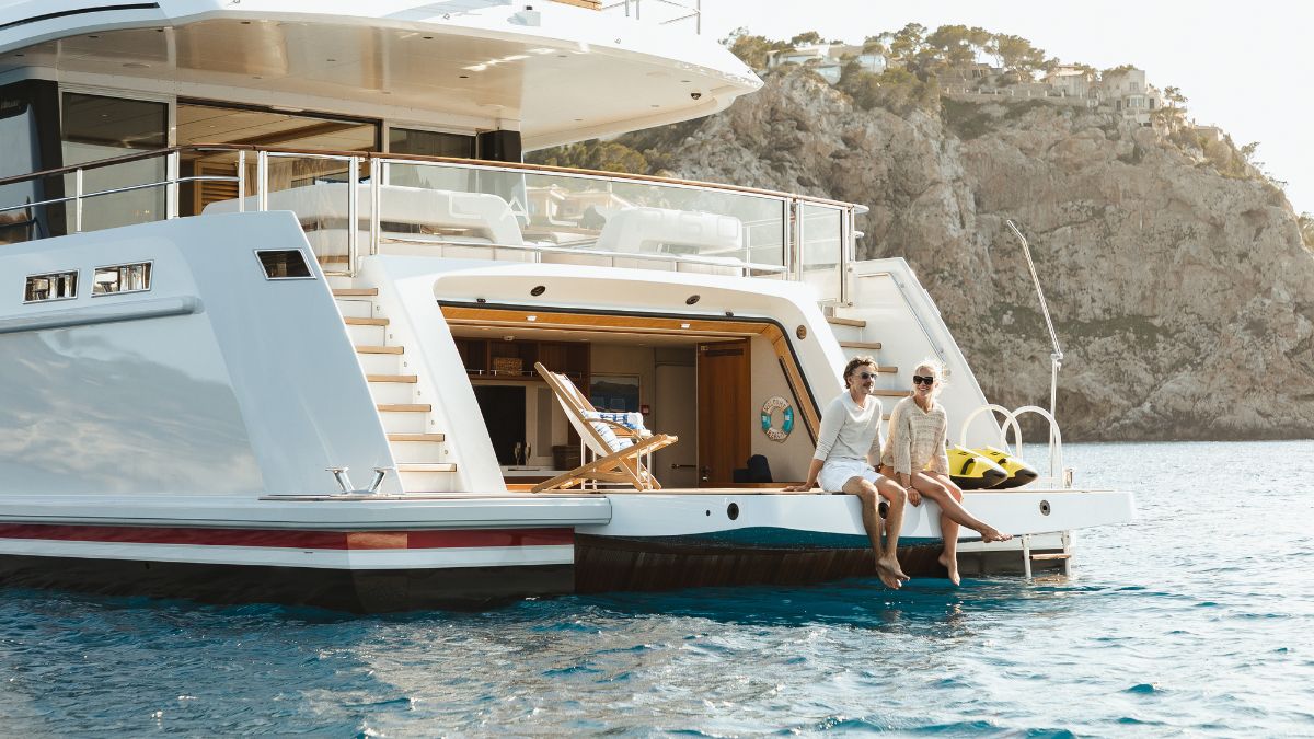 Two women sit on the stern of a luxurious yacht, dipping their feet in the turquoise water. The yacht's sleek design complements its stunning coastal backdrop with rocky cliffs and a sunny sky. A lounge chair and colorful water gear are visible nearby.