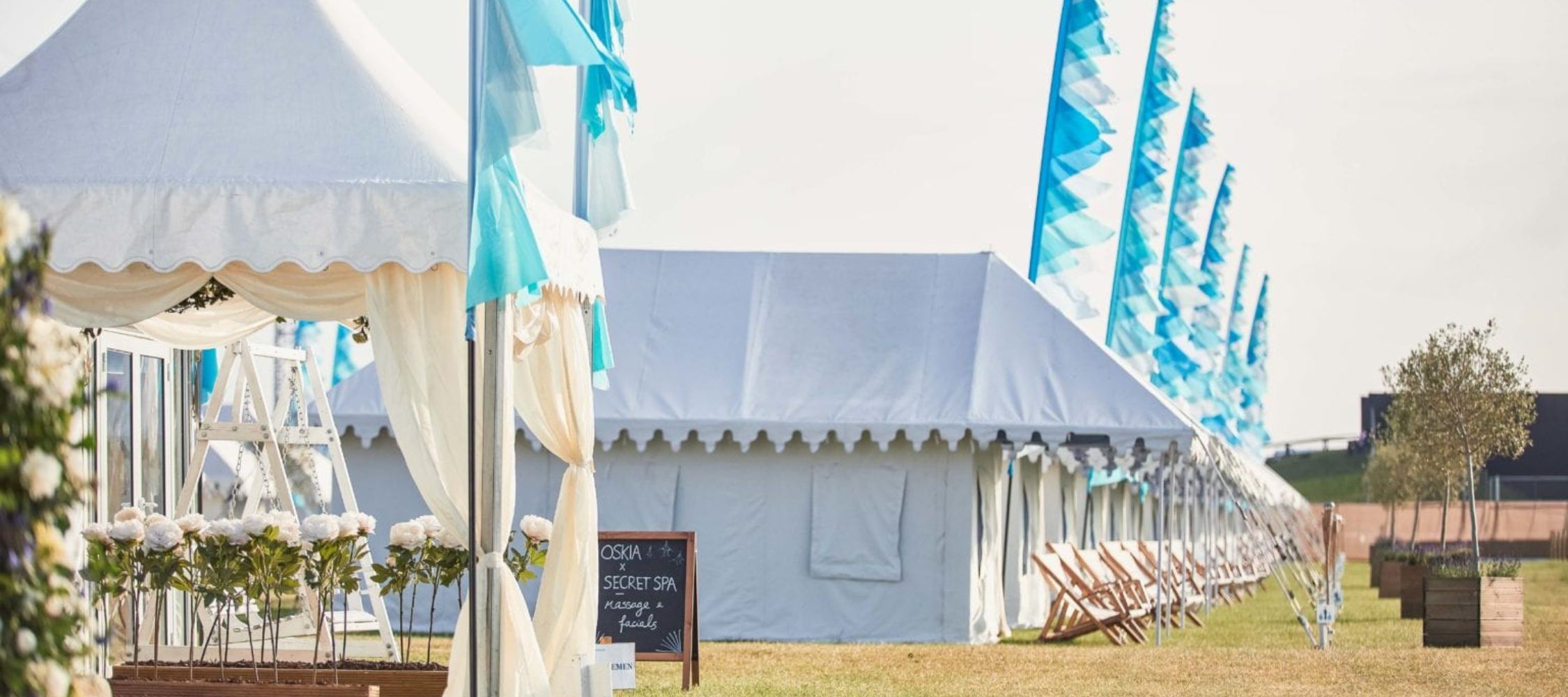 A row of large white tents with light blue accents is set up on a grassy field, decorated with floral arrangements. Colorful flags flutter in the breeze, and a wooden sign indicates a private event.