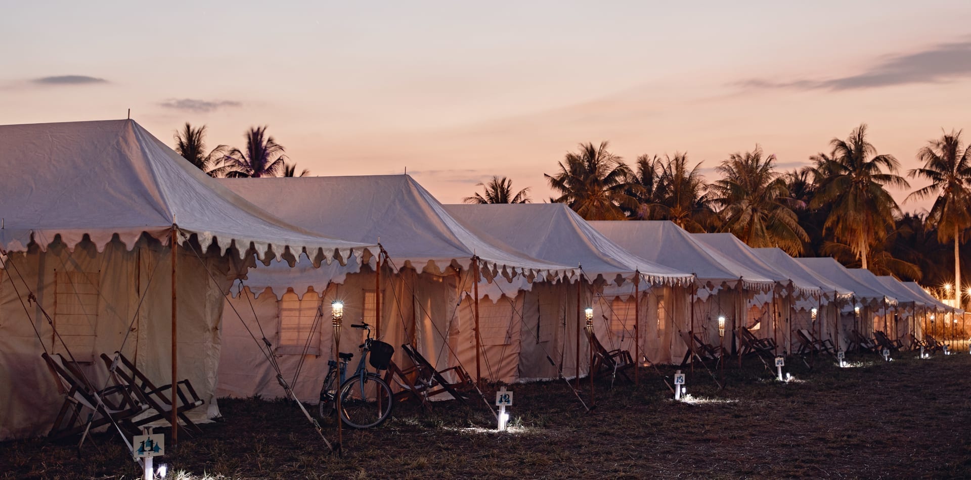 Row of white tents lined up on a grassy field at dusk, with palm trees in the background. Soft lights illuminate the area, and a bicycle is parked next to one of the tents.