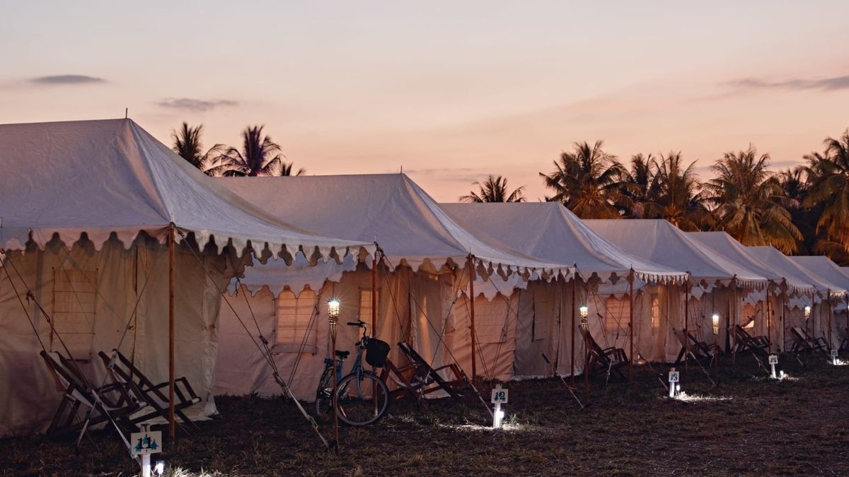 A row of white tents sits on a grassy field at sunset, with palm trees in the background. Soft lighting illuminates the tents, and there is a bicycle leaning against one.