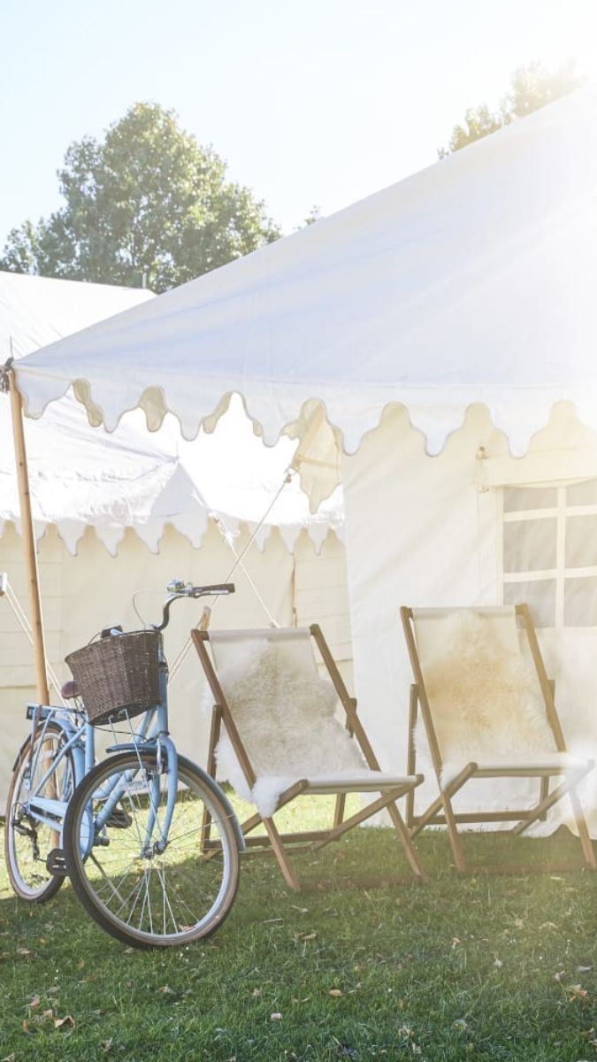A blue bicycle with a basket leans against two wooden deck chairs beside a white marquee tent on a sunny day.