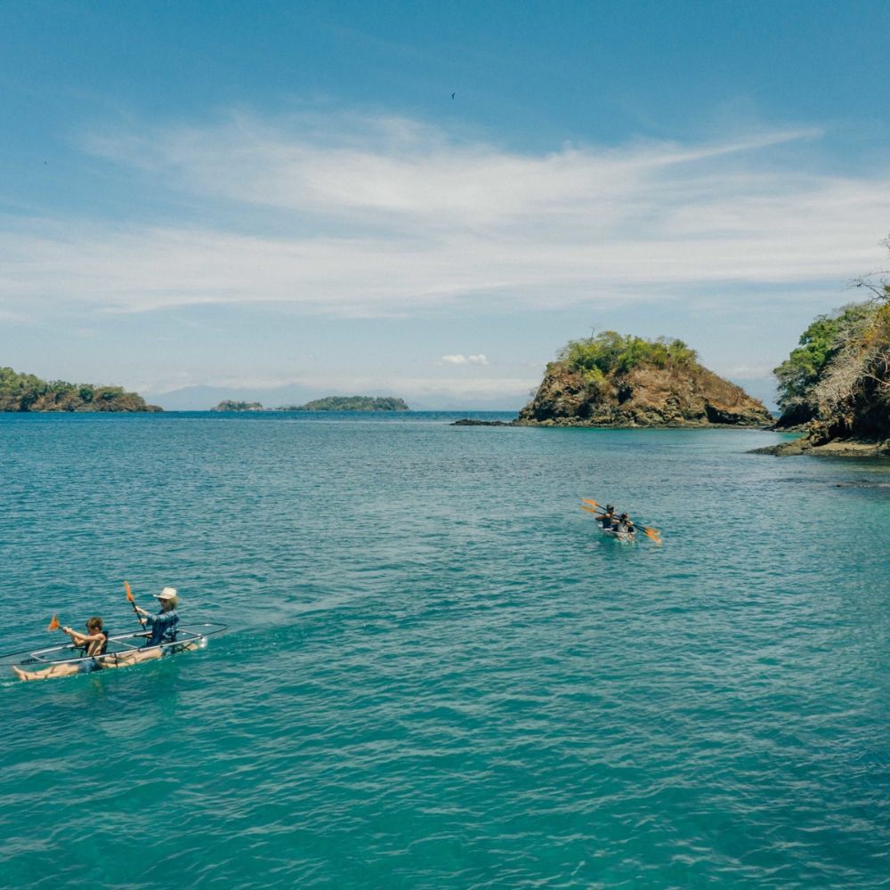 Two groups of people paddle canoes across calm blue waters, with lush green islands visible in the background under a clear sky.