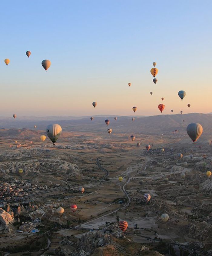 Colorful hot air balloons fill the sky above a picturesque landscape, with rolling hills and valleys illuminated by a soft sunrise. The scene captures the tranquil beauty of an early morning in a serene setting.