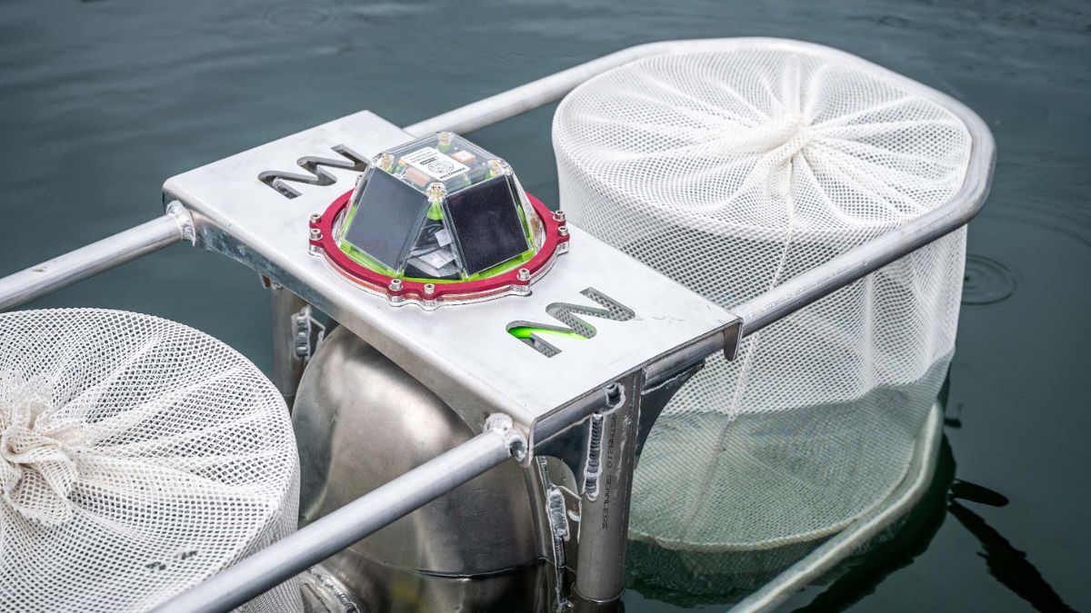 A metallic device used for water sampling, featuring two mesh nets on either side and a central sensor housing with multiple LED lights on top. The equipment is partially submerged in water.