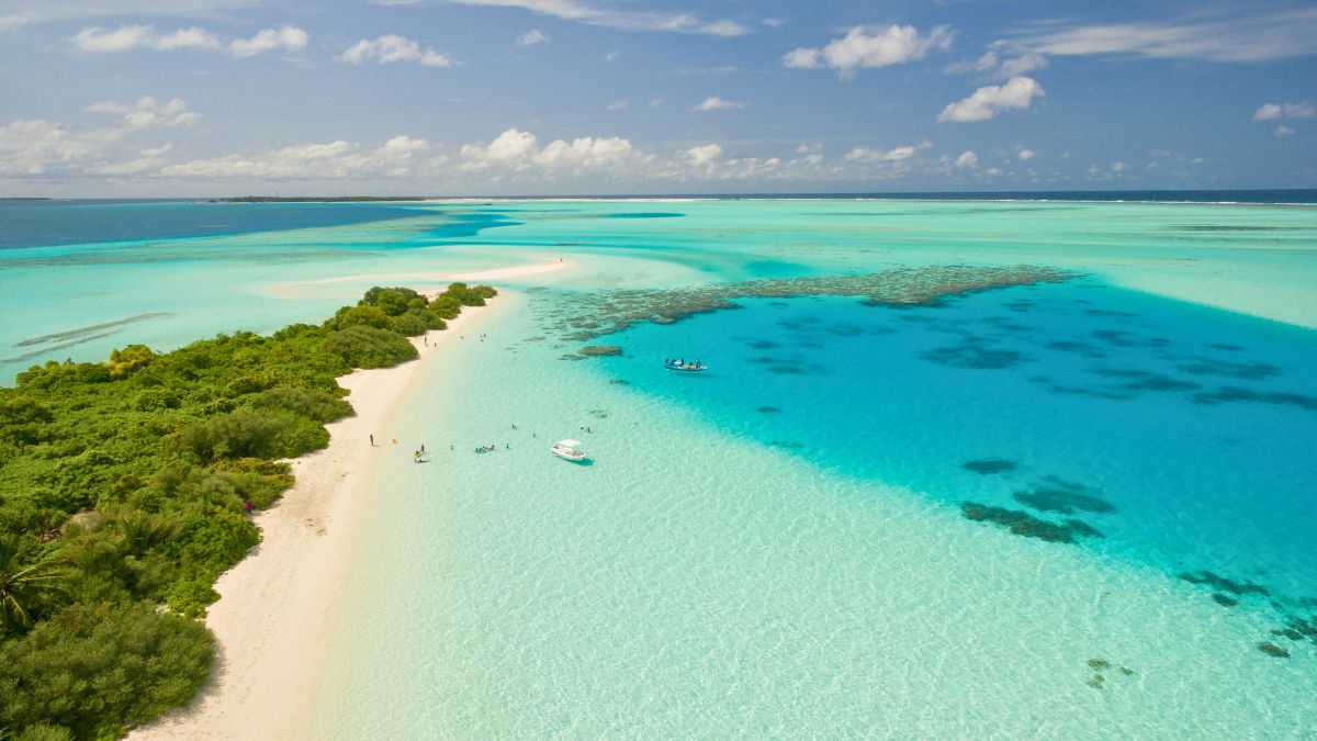 Aerial view of a stunning tropical lagoon with turquoise waters, sandy beaches, and lush green vegetation. Small boats are visible near the shore, surrounded by coral reefs. Fluffy white clouds drift across a bright blue sky.