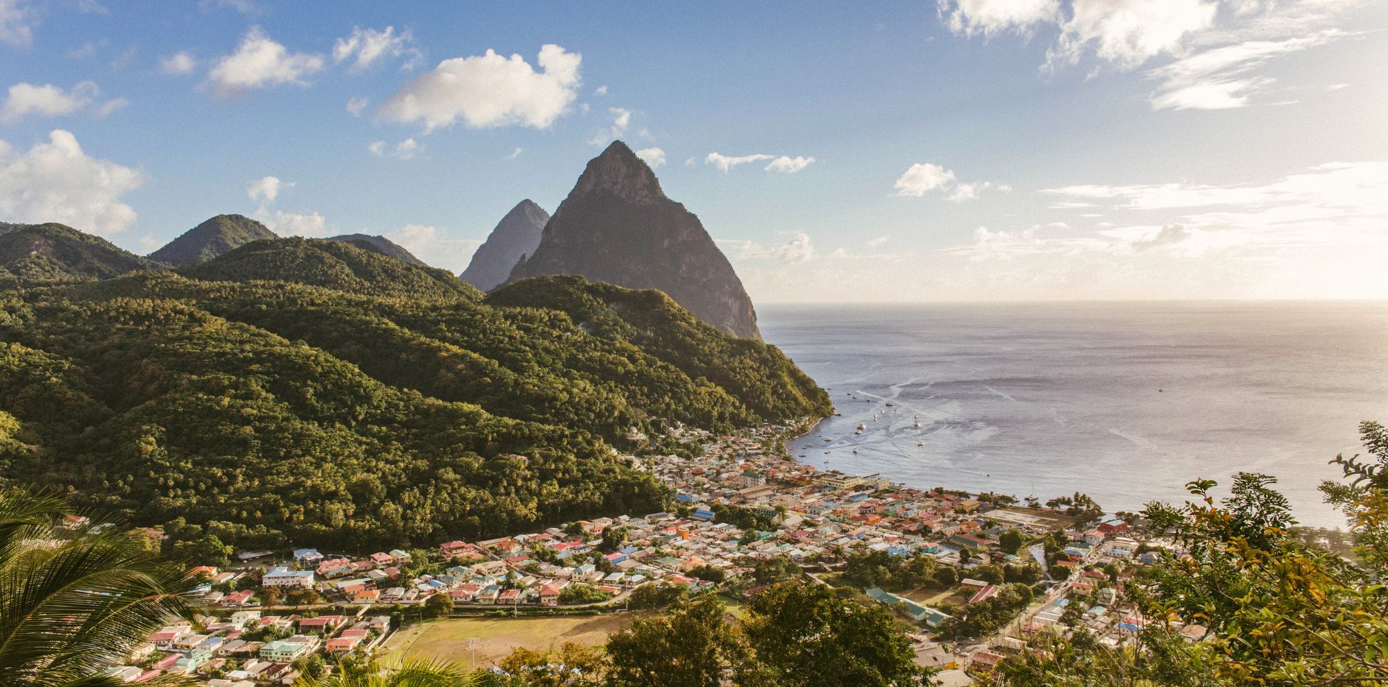 A lush coastal landscape features a vibrant town with colorful buildings nestled at the foot of towering green hills. The iconic Pitons rise majestically from the sea under a bright blue sky with scattered clouds.