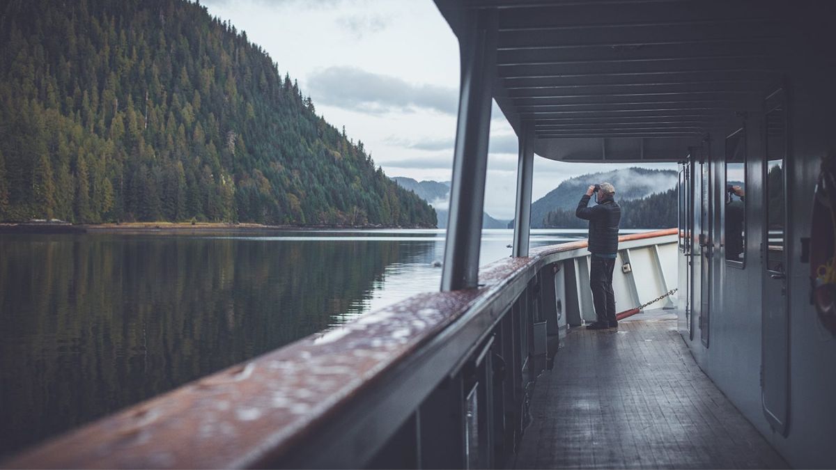 A person stands on the deck of a boat, gazing out at a calm river surrounded by lush green mountains and trees. The water reflects the scenery, creating a serene atmosphere.