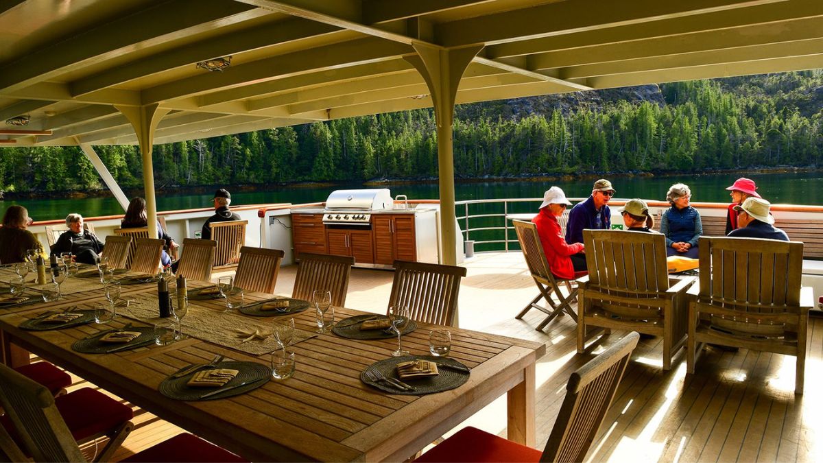 A spacious outdoor dining area on a boat, featuring a large wooden table set for a meal with glassware and plates. In the background, several people are seated at a smaller table enjoying conversation, surrounded by lush green trees and calm water. A barbecue grill is positioned near the dining area.