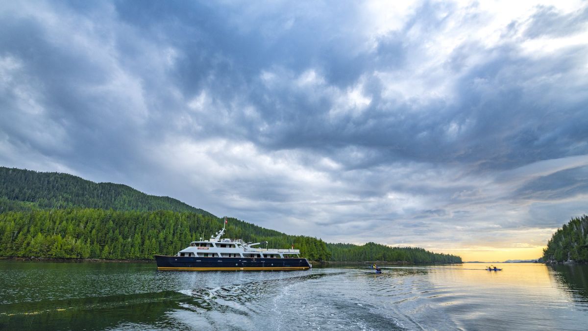 A large vessel navigates a calm waterway surrounded by dense green forests and dramatic cloudy skies. Smaller kayakers paddle nearby, reflecting the scenic landscape.