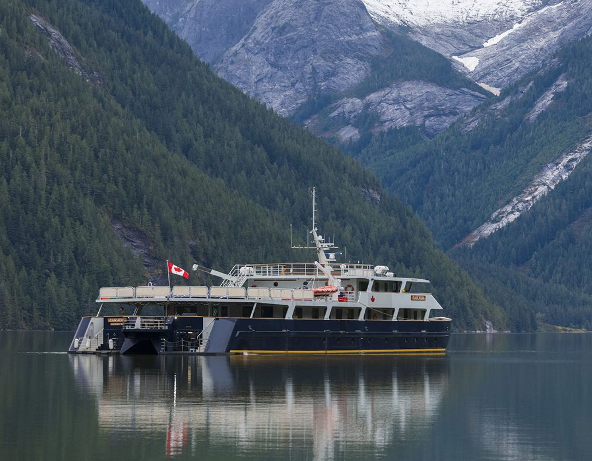 A large ferry boat sits anchored in a calm, reflective body of water, surrounded by steep mountains covered in lush green forest. Snow-capped peaks are visible in the background, hinting at a rugged landscape. The boat features a Canadian flag on the side.